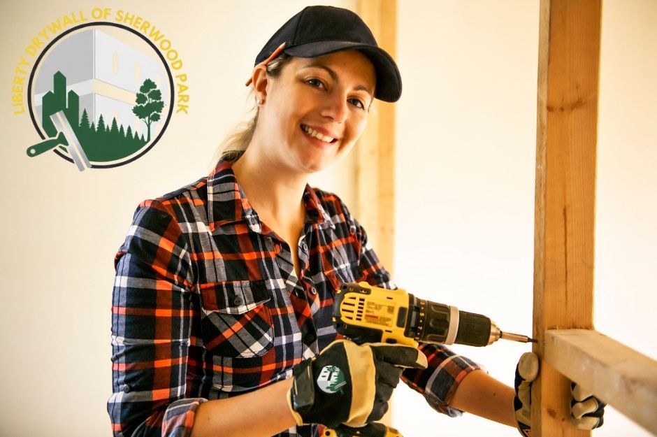 A drywall repair specialist smiles while operating a yellow drill during a residential renovation project. She wears a black cap and plaid flannel shirt, working among exposed wooden framing near Osborne Dr, and Range Rd 264, Acheson, AB.