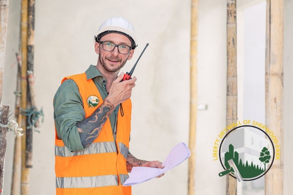 A drywall repair specialist wearing a white hard hat, glasses, and an orange safety vest is standing inside a residential construction site. He holds a red walkie-talkie in one hand and blueprints in the other, surrounded by bamboo scaffolding. Located near Hillcrest Point, and Greenfield Way, Fort Saskatchewan, AB.