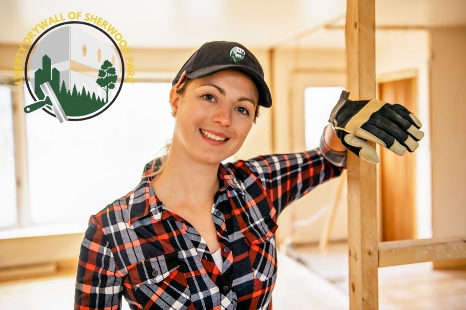A drywall repair specialist in a plaid shirt and black cap smiles while leaning against wooden framing in a commercial construction space with bright lighting and structural beams visible near Chippewa Rd, and Corinthia Dr, Leduc, AB.