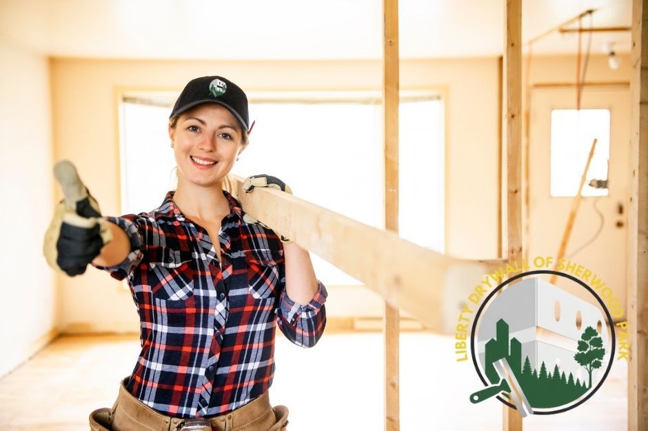 A drywall repair technician in a plaid shirt and black cap smiles while holding wood in a residential construction space with wooden framing and bright interior lighting visible near Kawartha St, and River Dr, Devon, AB.