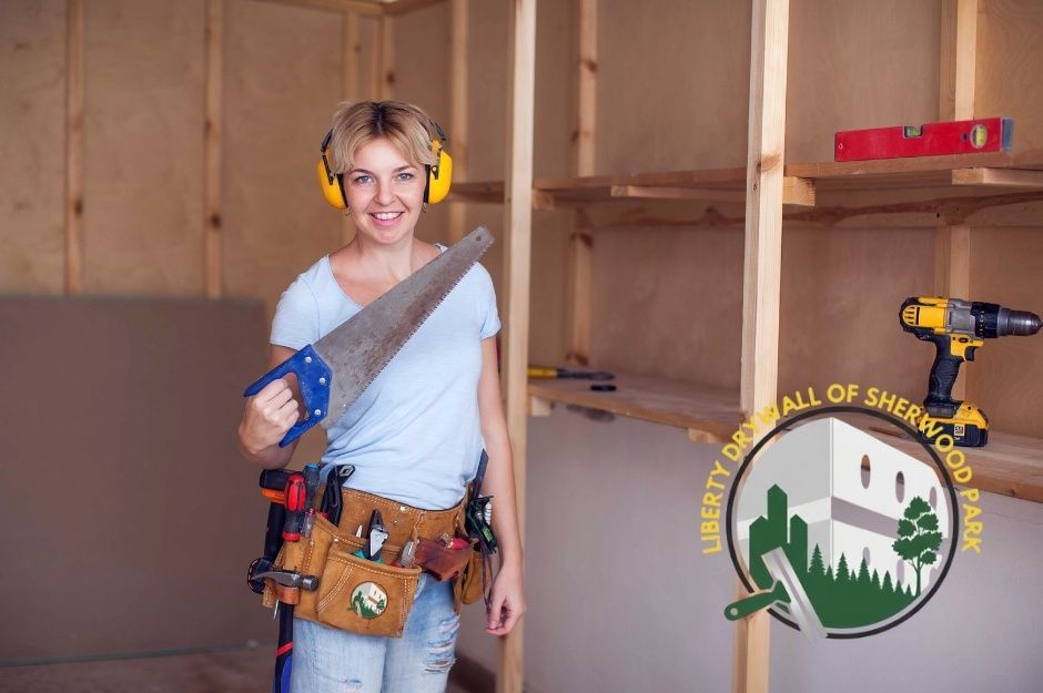 A drywall repair technician wearing yellow ear protection and a tool belt smiles while holding a saw in a commercial construction space with exposed wooden framing and tools visible near 8 St, and 25 Ave, Nisku, AB.