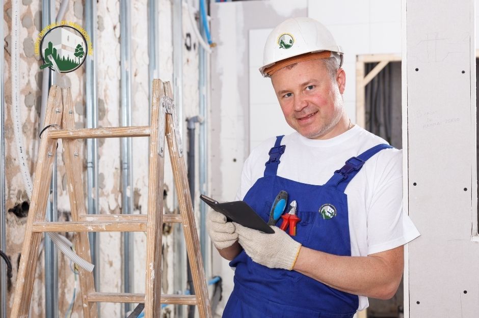 A photo of a man doing drywall services, wearing blue overalls and a hard hat, smiling while holding a clipboard and pen beside a wooden ladder and exposed metal wall studs with visible wiring inside an unfinished residential room in Sherwood Park, AB.