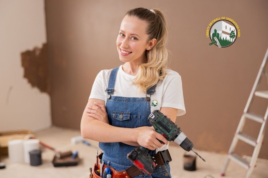 An employee of Liberty Drywall of Sherwood Park smiling while holding a cordless drill, wearing denim overalls and a tool belt, standing in a room with partially painted walls, paint rollers, and a ladder during drywall repair and finishing in Sherwood Park, AB.