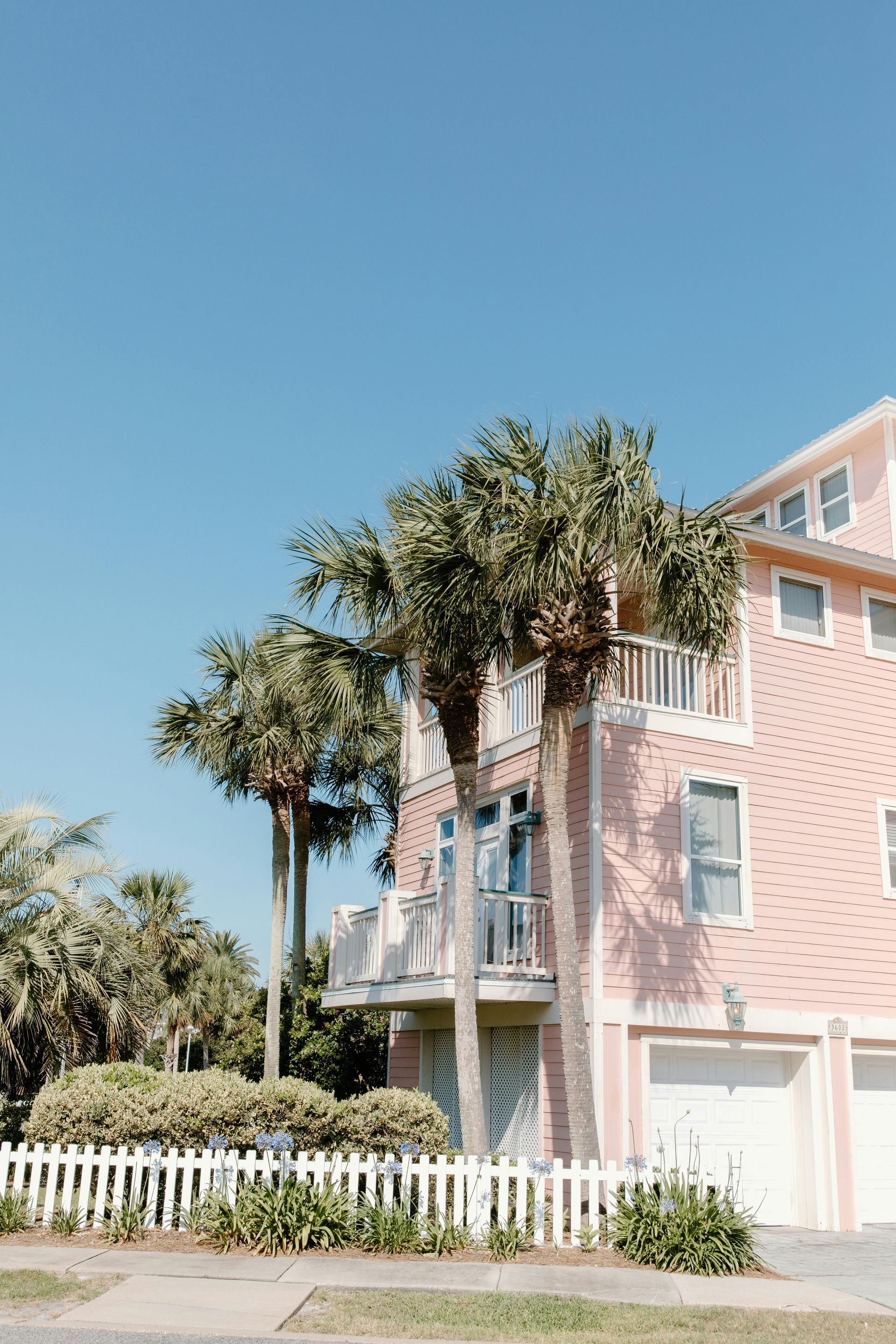 Pink beach house with palm trees, white fence, and blue sky.