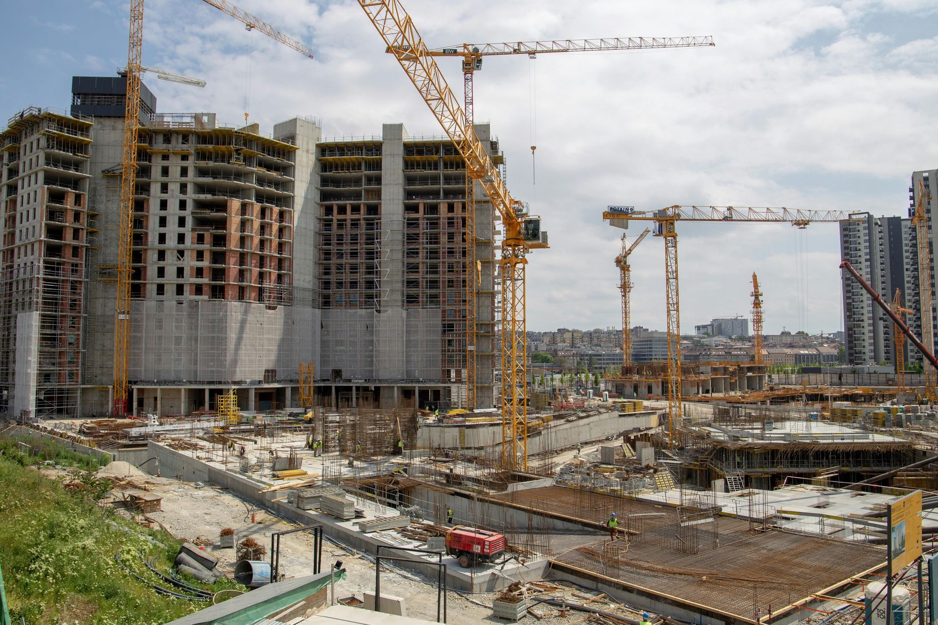 Large construction site with cranes, unfinished buildings, and excavated ground under a cloudy sky