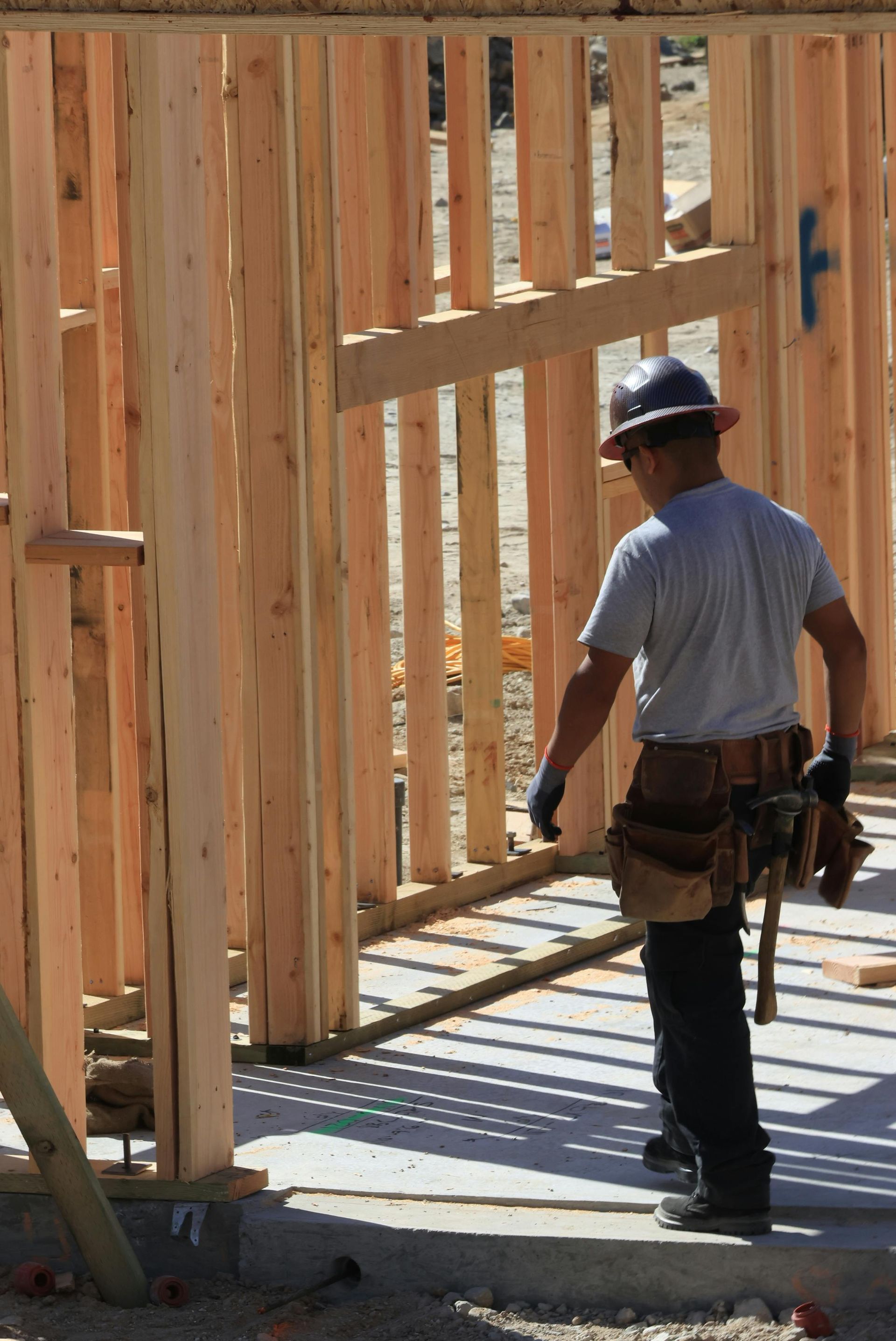 Construction worker at a wooden house frame, wearing a hard hat and tool belt, inspecting the site