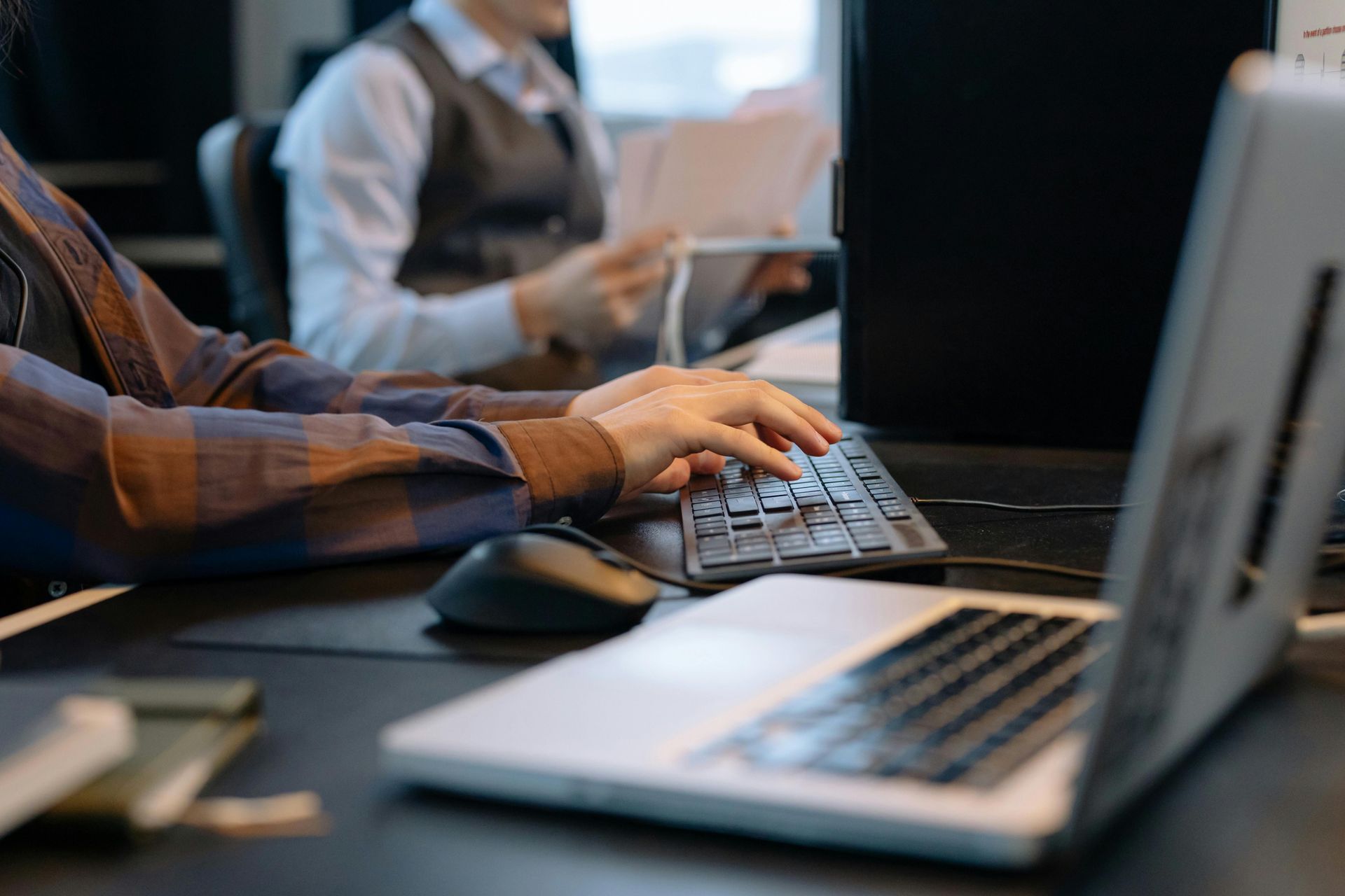 Hands typing on a keyboard beside a laptop in an office setting, with people blurred in the background
