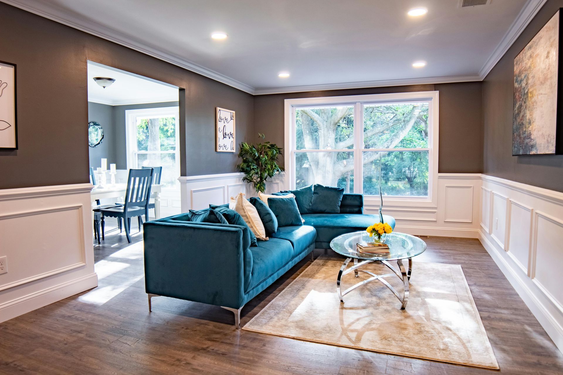 Bright living room with teal sofa, glass coffee table, beige rug, and large windows overlooking greenery