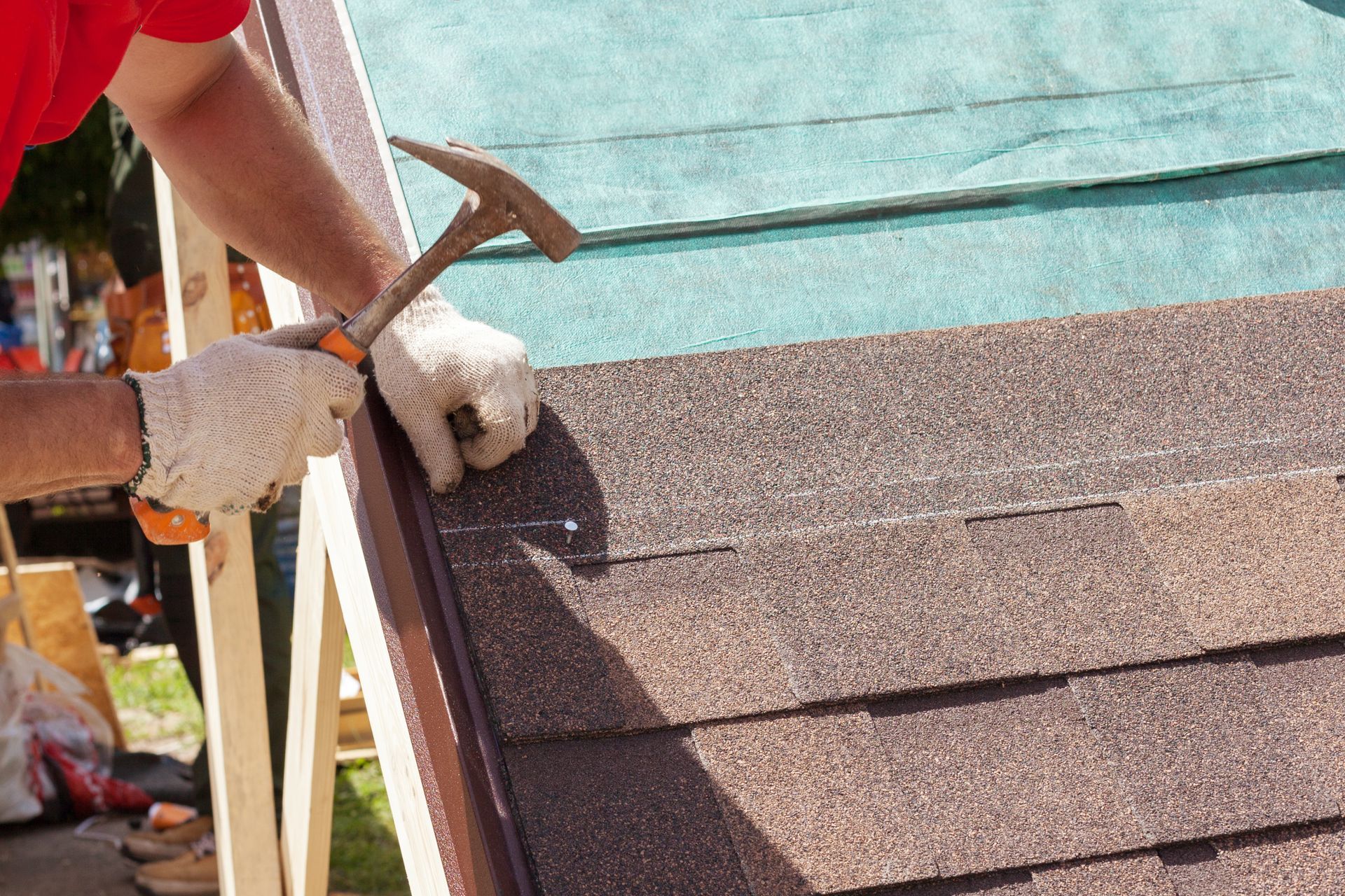 Roofing worker hammering asphalt shingles, wearing gloves and red shirt on residential rooftop.