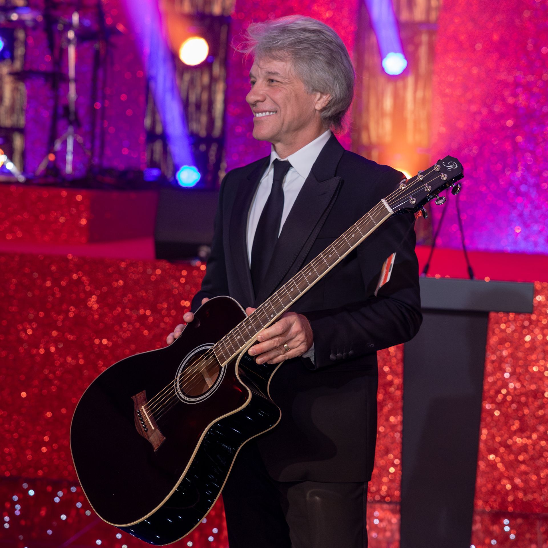 Musician Jon Bon Jovi performs at a corporate event in London, captured by a professional photographer at Ghelani Studios. The photo shows Jon Bon Jovi on stage, holding a guitar, smiling, and looking out toward the audience.