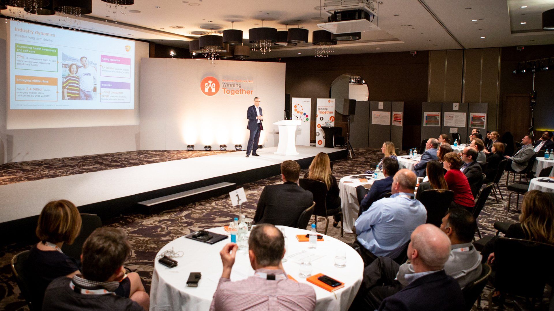 Business professionals networking at a corporate event. The attendees are standing in front of a backdrop displaying event branding, with drinks and refreshments on a table in the foreground. Photo taken by Ghelani Studios, professional event photographers in London and the UK.