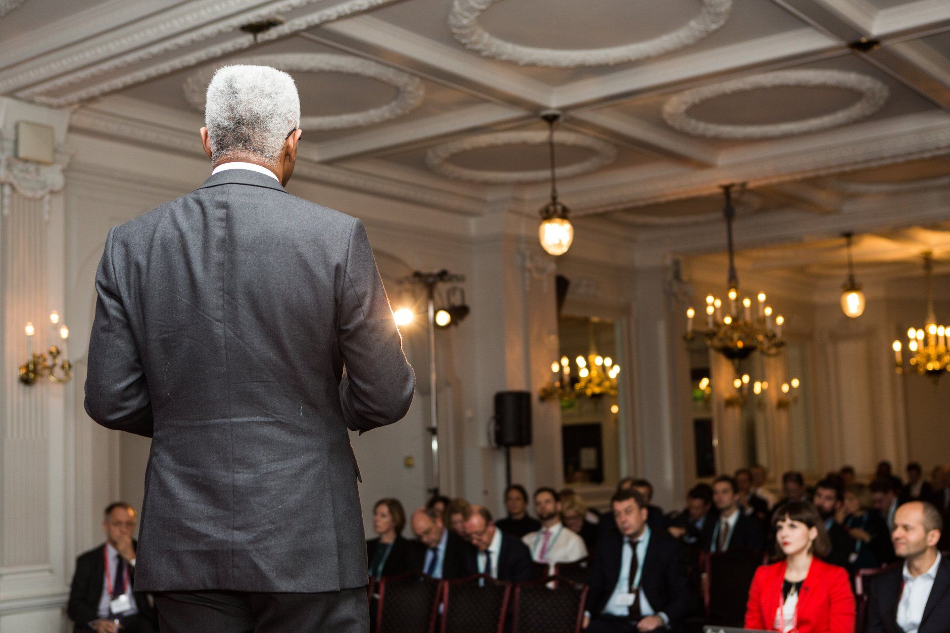 Back of keynote speaker standing on stage. The conference attendees are seated in rows facing a stage, with a large screen displaying event branding behind the stage. The aisle is spacious and filled with natural light. Photo taken by Ghelani Studios, experienced conference photographers in London.