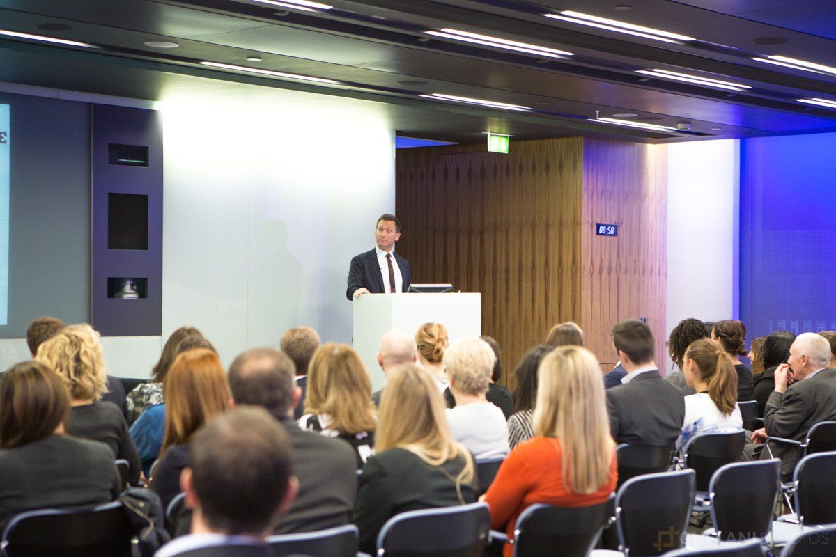 Medium shot photograph of a conference aisle. The conference attendees are seated in rows facing a stage, with a large screen displaying event branding behind the stage. The aisle is spacious and filled with natural light. Photo taken by Ghelani Studios, experienced conference photographers in London.