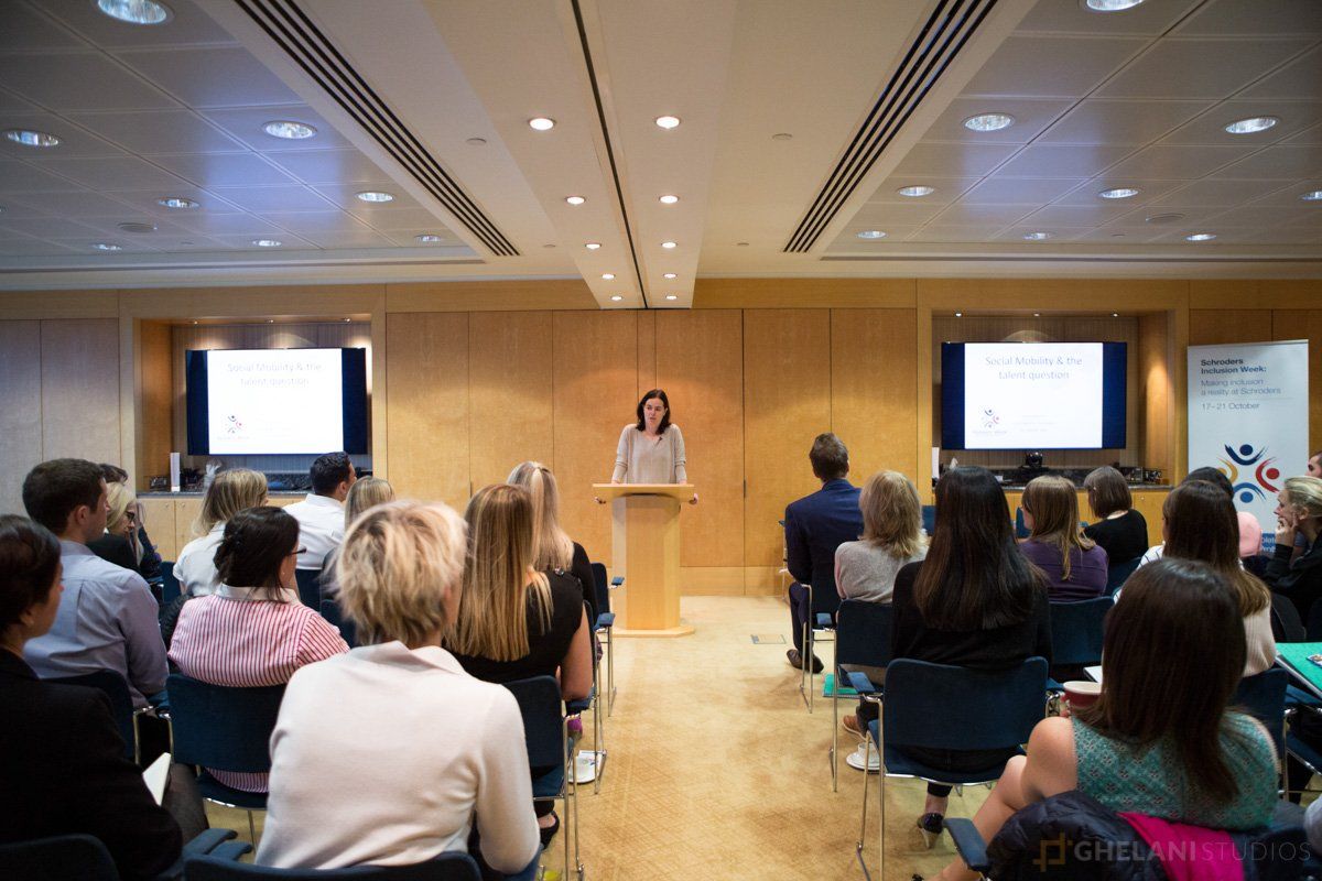 Wide shot photograph of a conference aisle. The conference attendees are seated in rows facing a stage, with a large screen displaying event branding behind the stage. The aisle is spacious and filled with natural light. Photo taken by Ghelani Studios, experienced conference photographers in London.