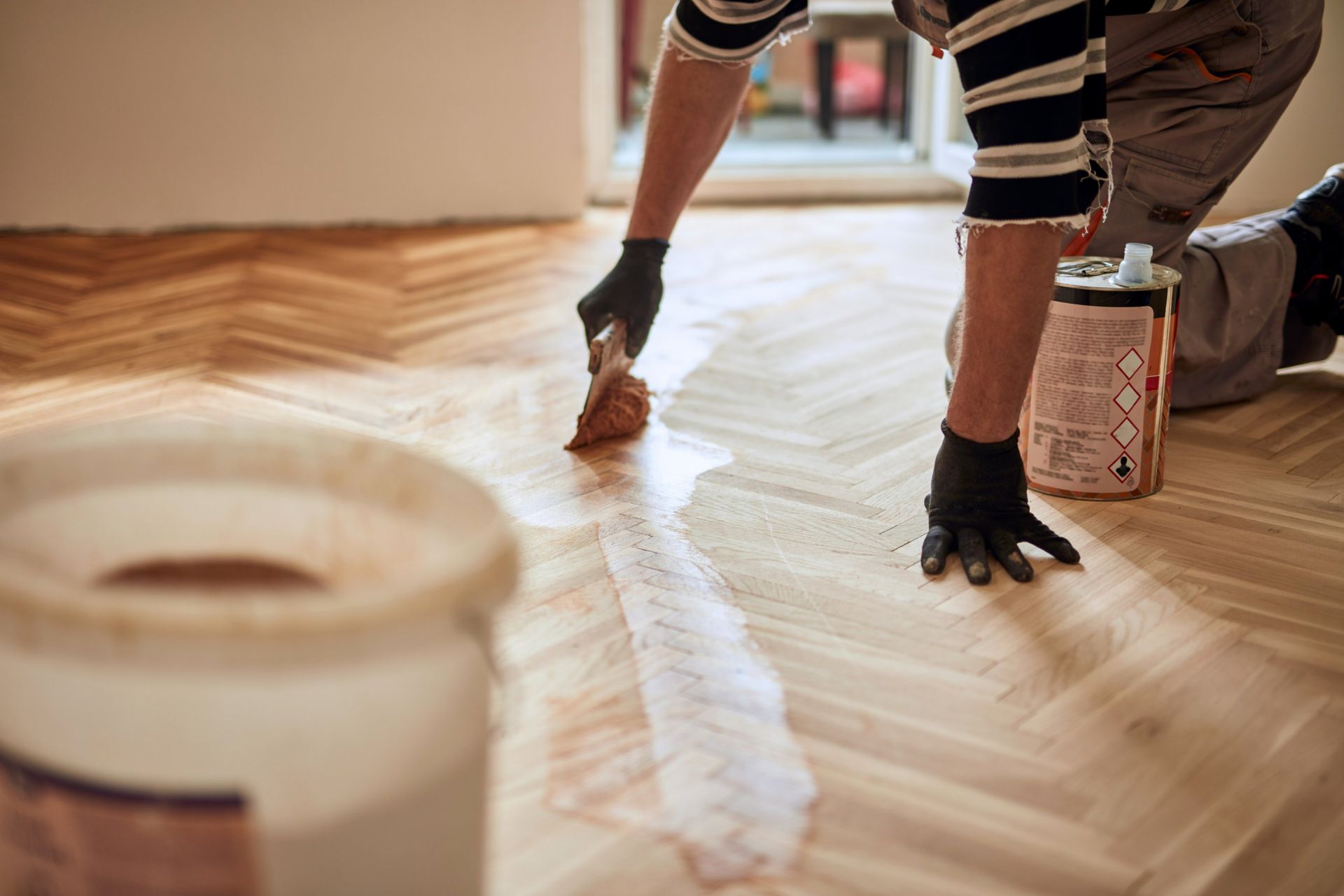 Person applies wood stain to parquet flooring with a brush, wearing gloves.