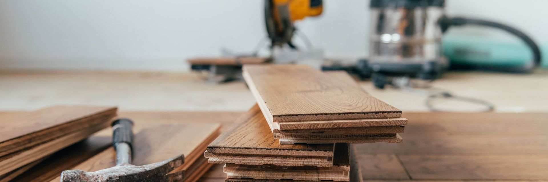 Wood planks stacked on a wooden surface, with tools like a saw in the background, possibly for construction.
