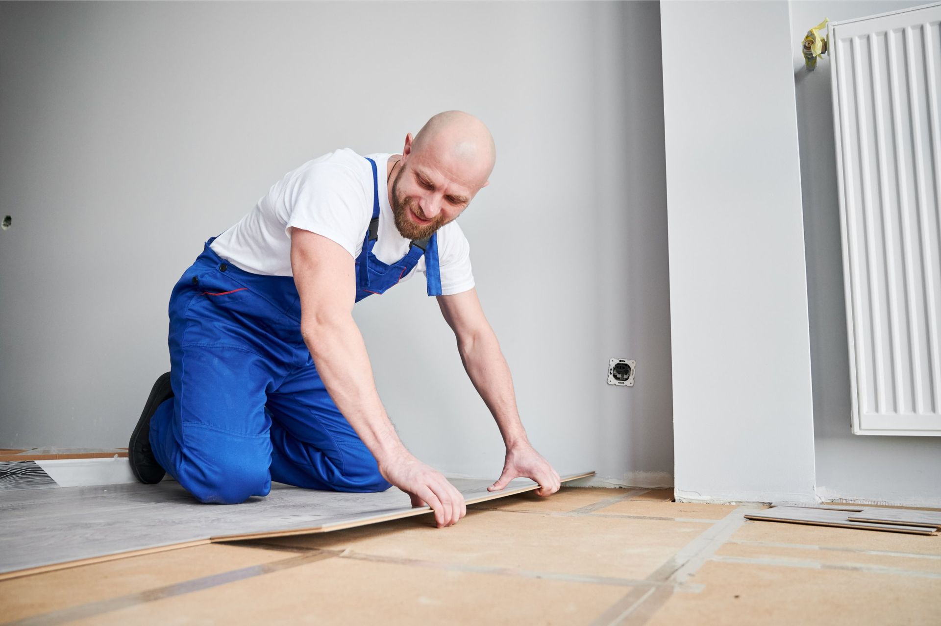 Man kneeling, installing flooring in a room with light gray walls and a radiator.
