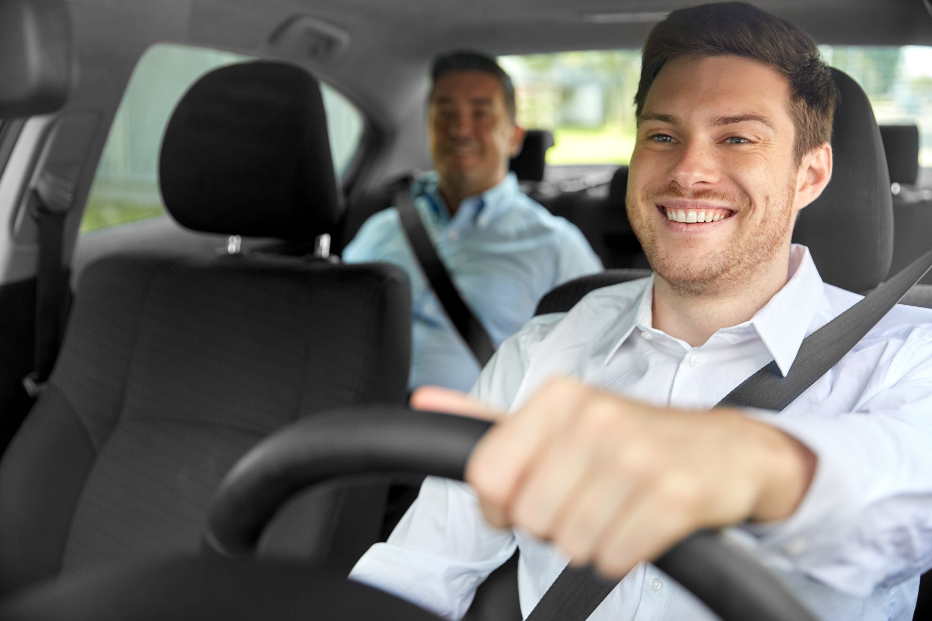 A man is driving a car with another man in the back seat.