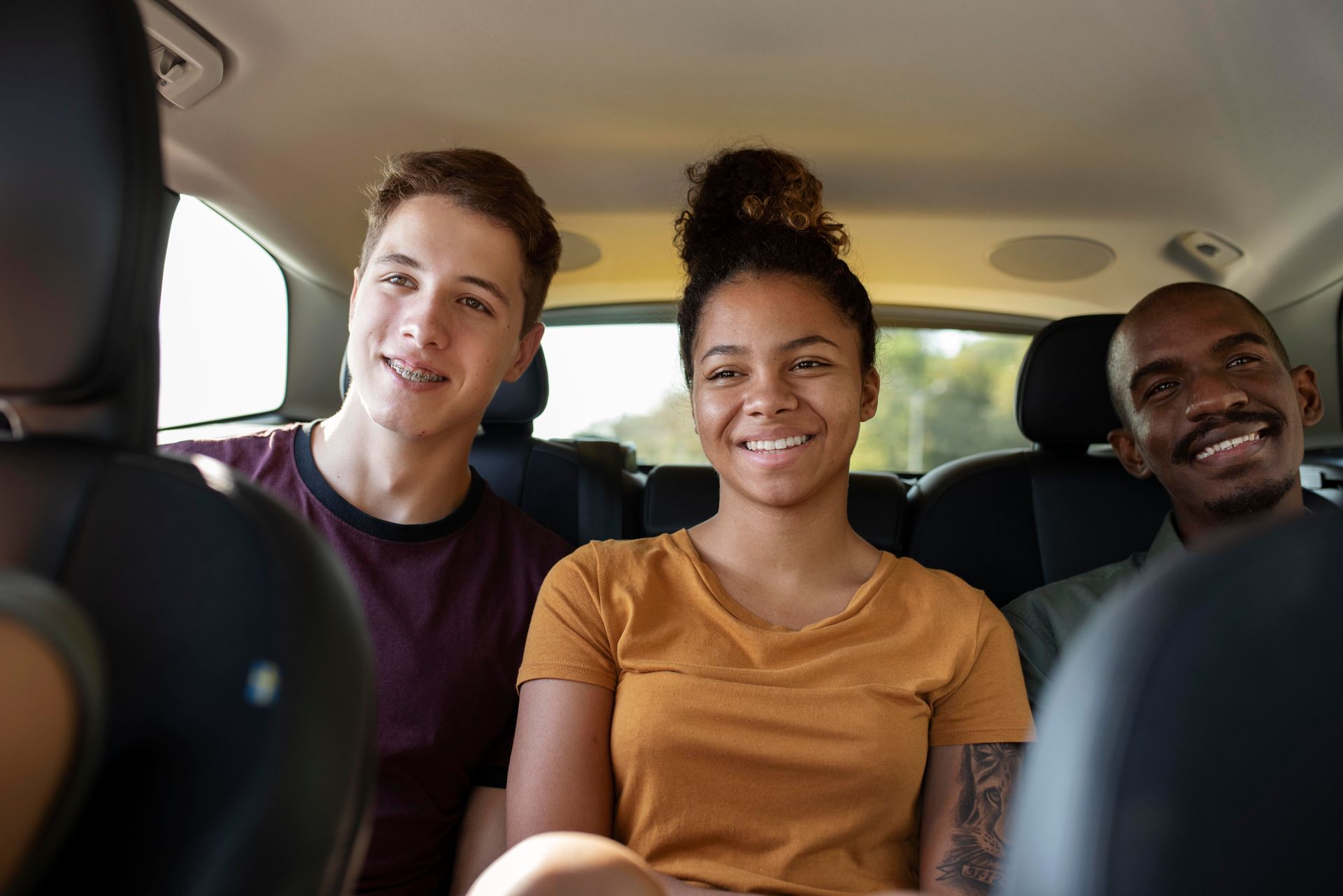 Three people smiling inside a car, two in the back seat. Sunlight.