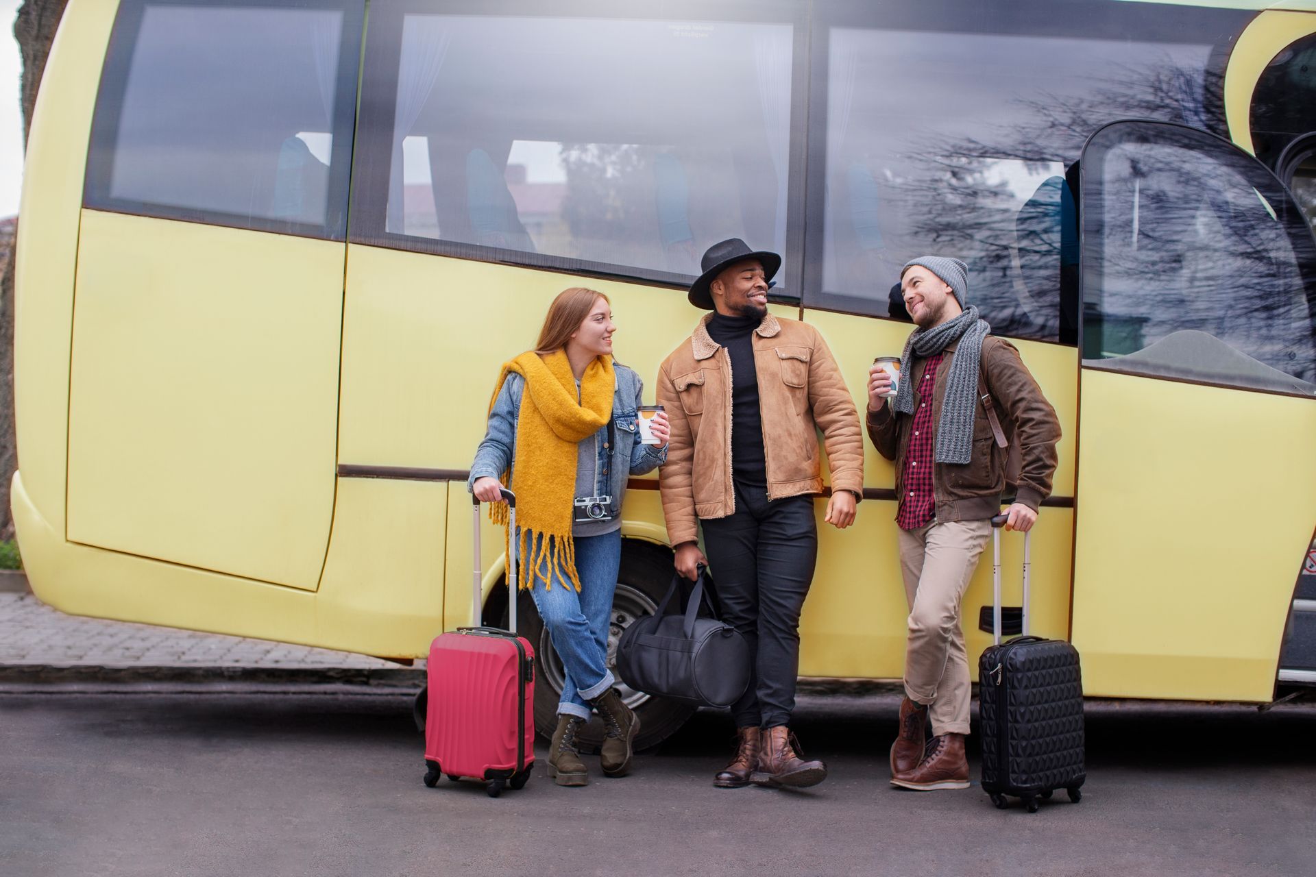 Three people with luggage stand near a yellow bus, conversing. One holds a coffee cup.