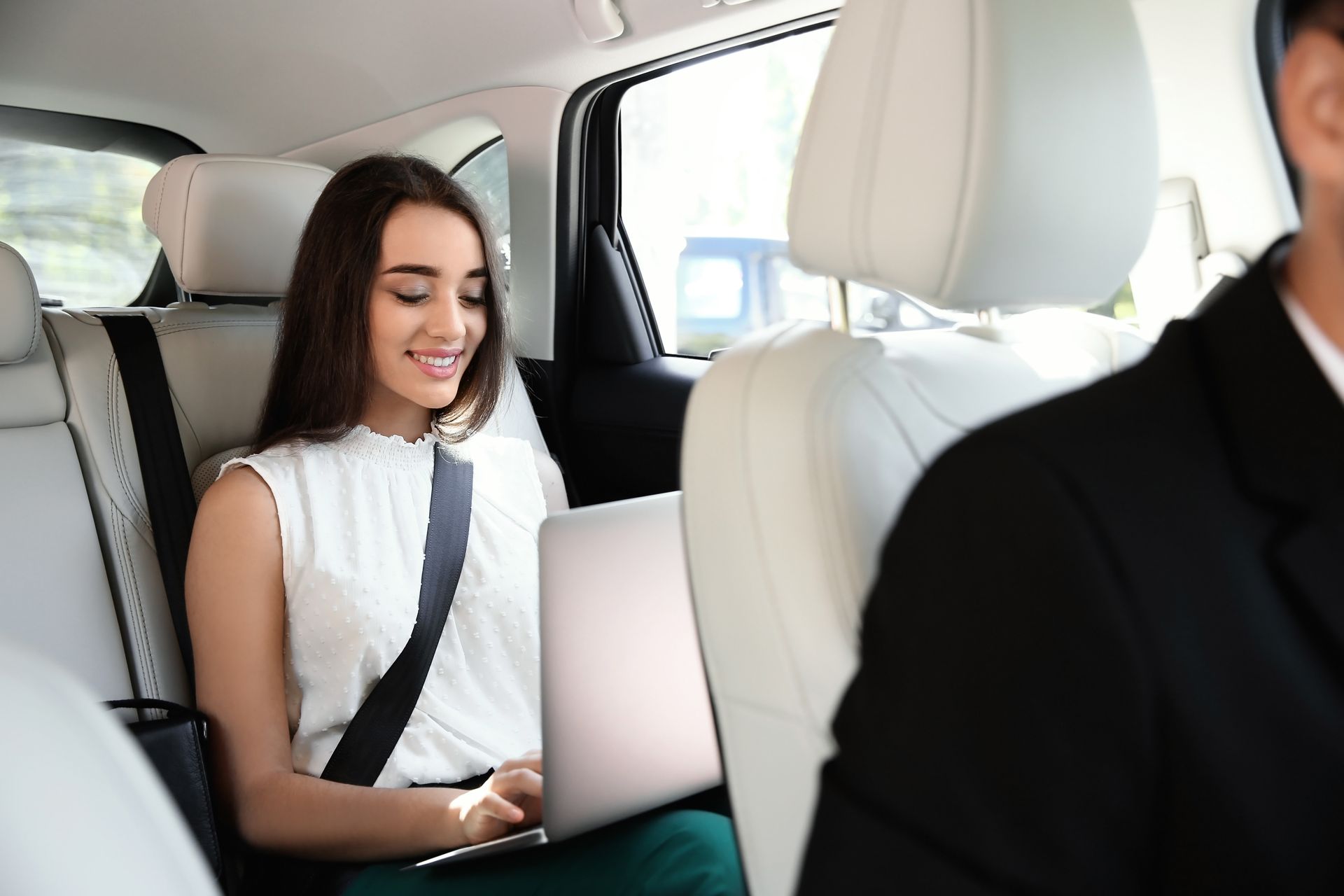 A woman is sitting in the back seat of a car using a laptop computer.
