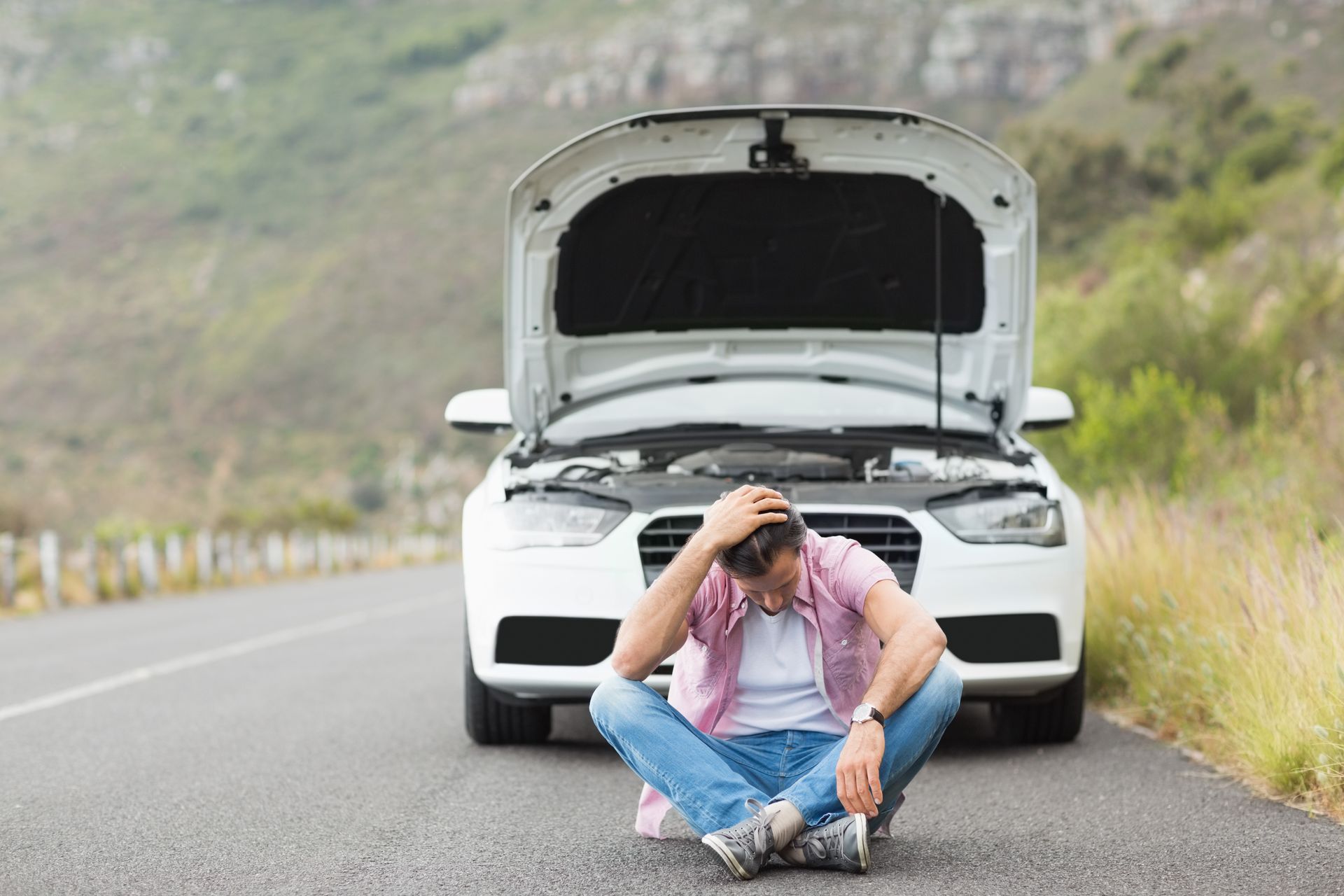 Man with head in hands sits in front of a broken-down white car on a road with mountains.
