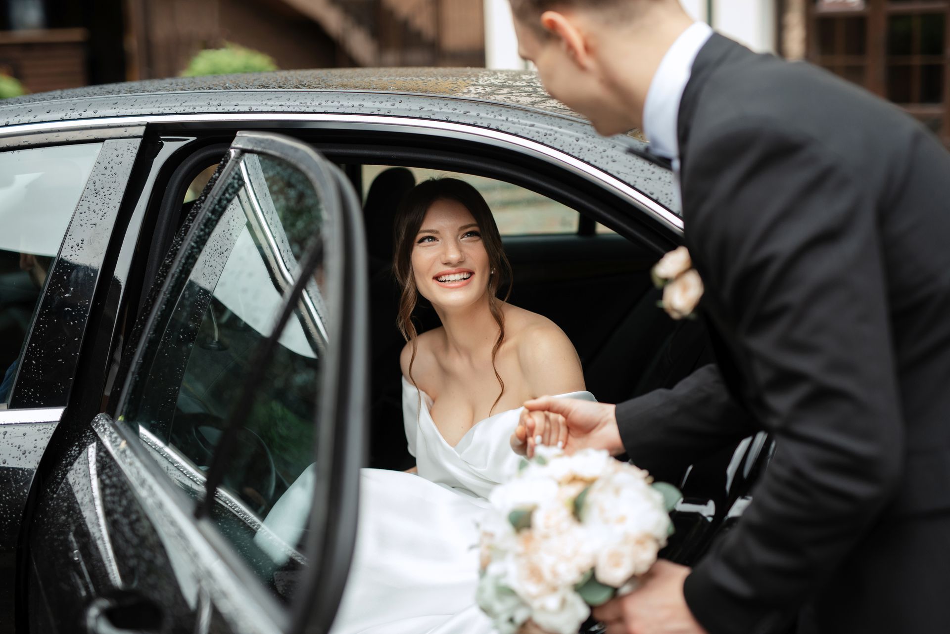 Bride exits a black car as groom helps her, holding bouquet. They smile in a rainy outdoor setting.