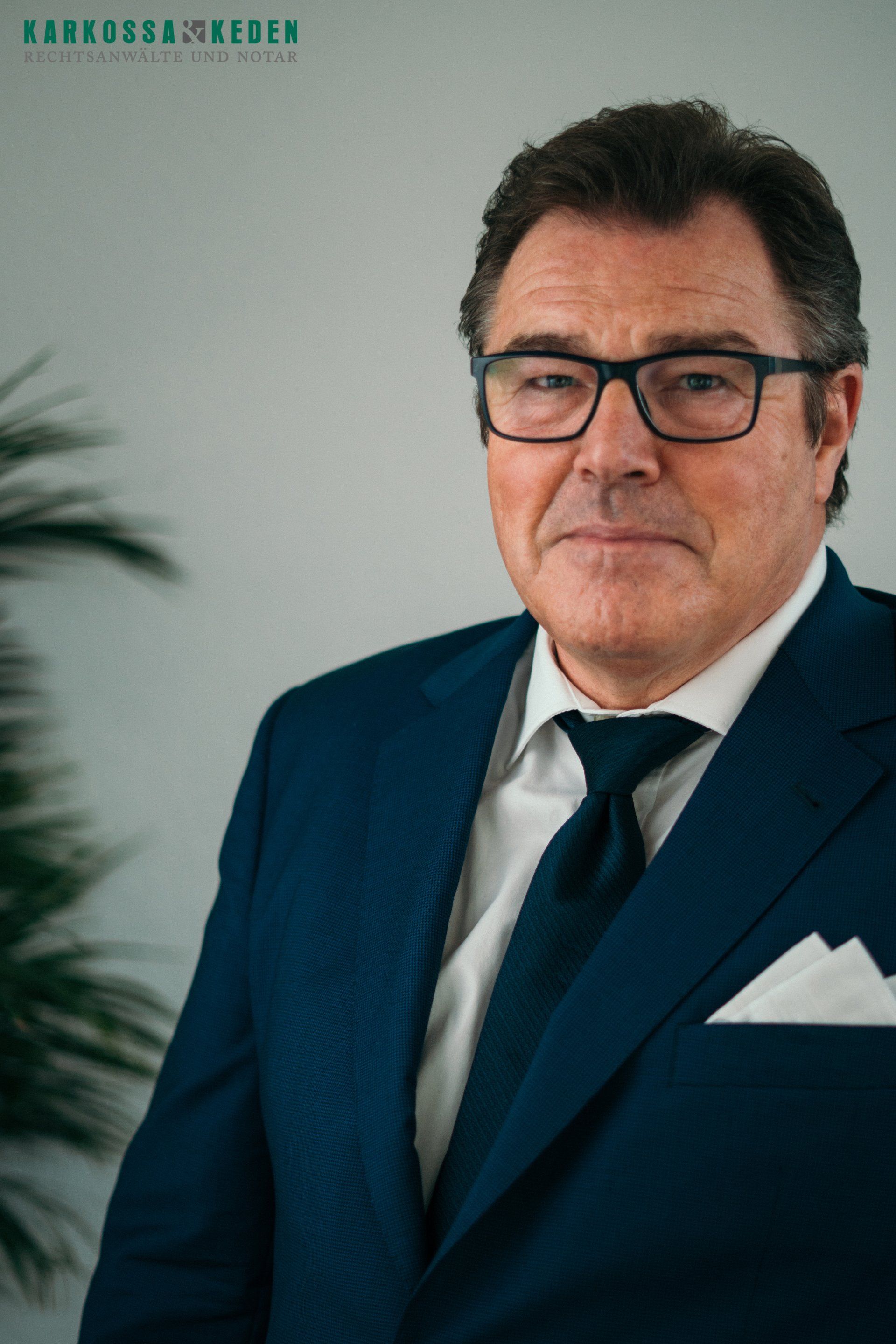 Mann in blauem Anzug und Brille blickt in die Kamera. Weißes Einstecktuch, neutraler Hintergrund.