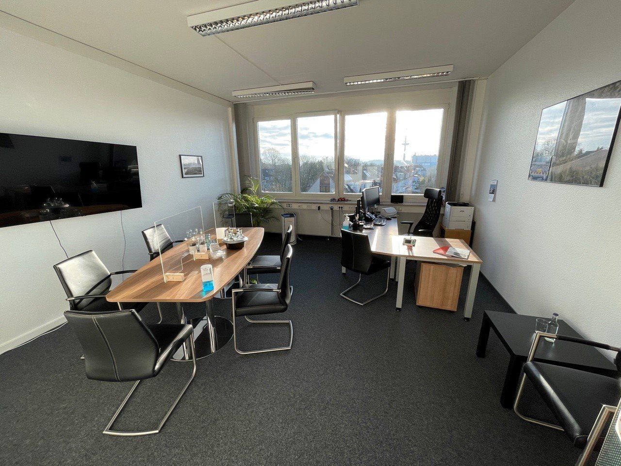 Ein Büro mit einem Konferenztisch, Stühlen und einem Schreibtisch, Fenstern mit Sonnenlicht und einem dunkelgrauen Teppich.
