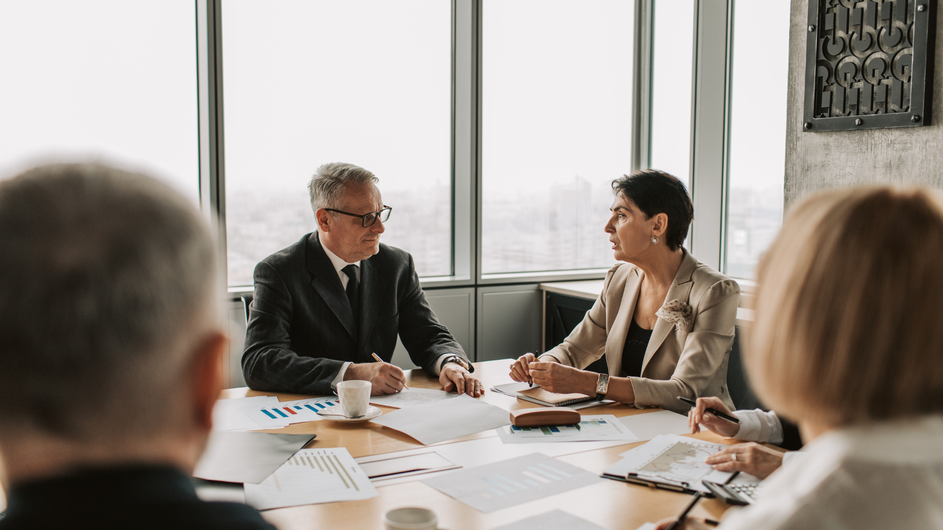 People in business attire at a conference table with documents, discussing. Window background.