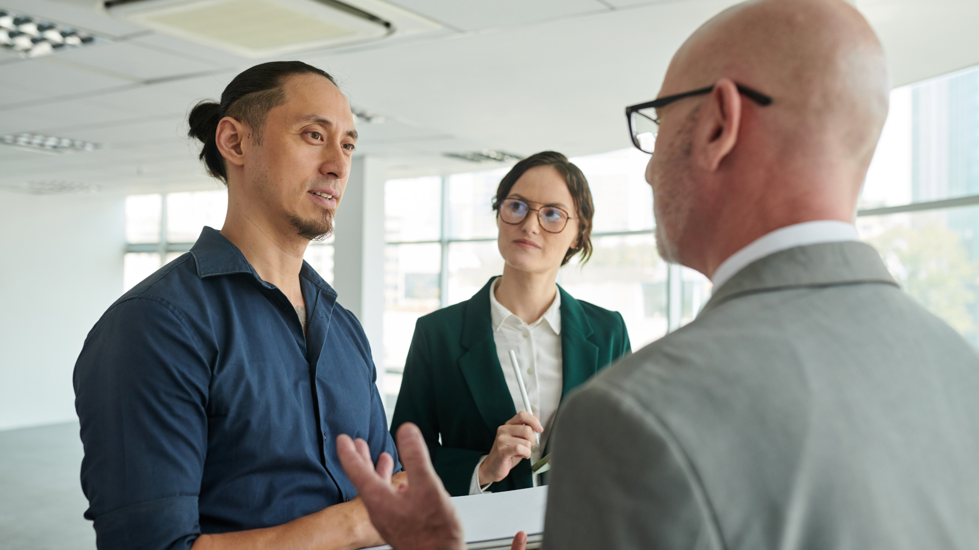 Three people in a bright office space; two conversing, one with arms gesturing, another watching attentively.
