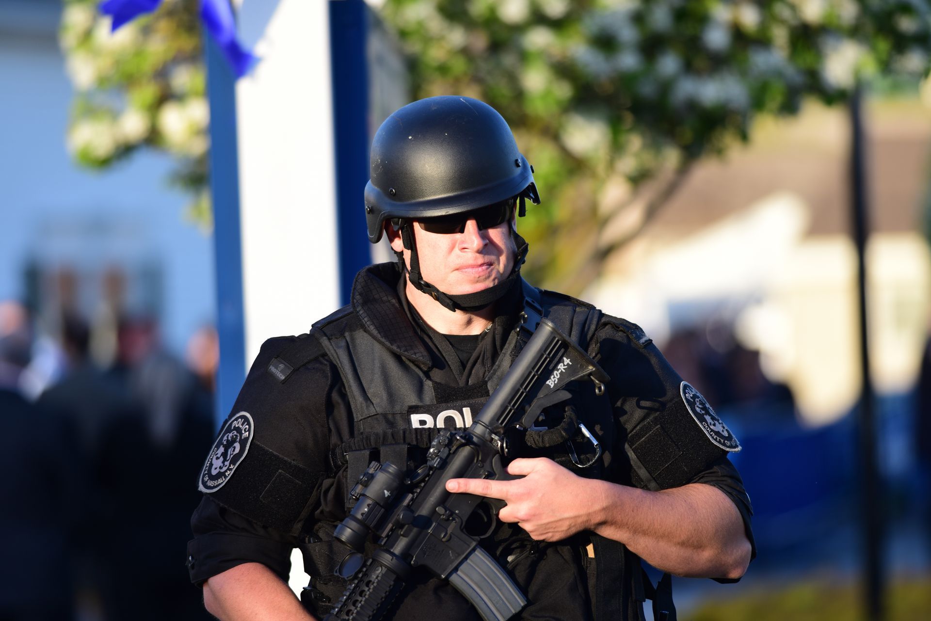 Armored police officer in black tactical gear holding a rifle outdoors near a blurred crowd