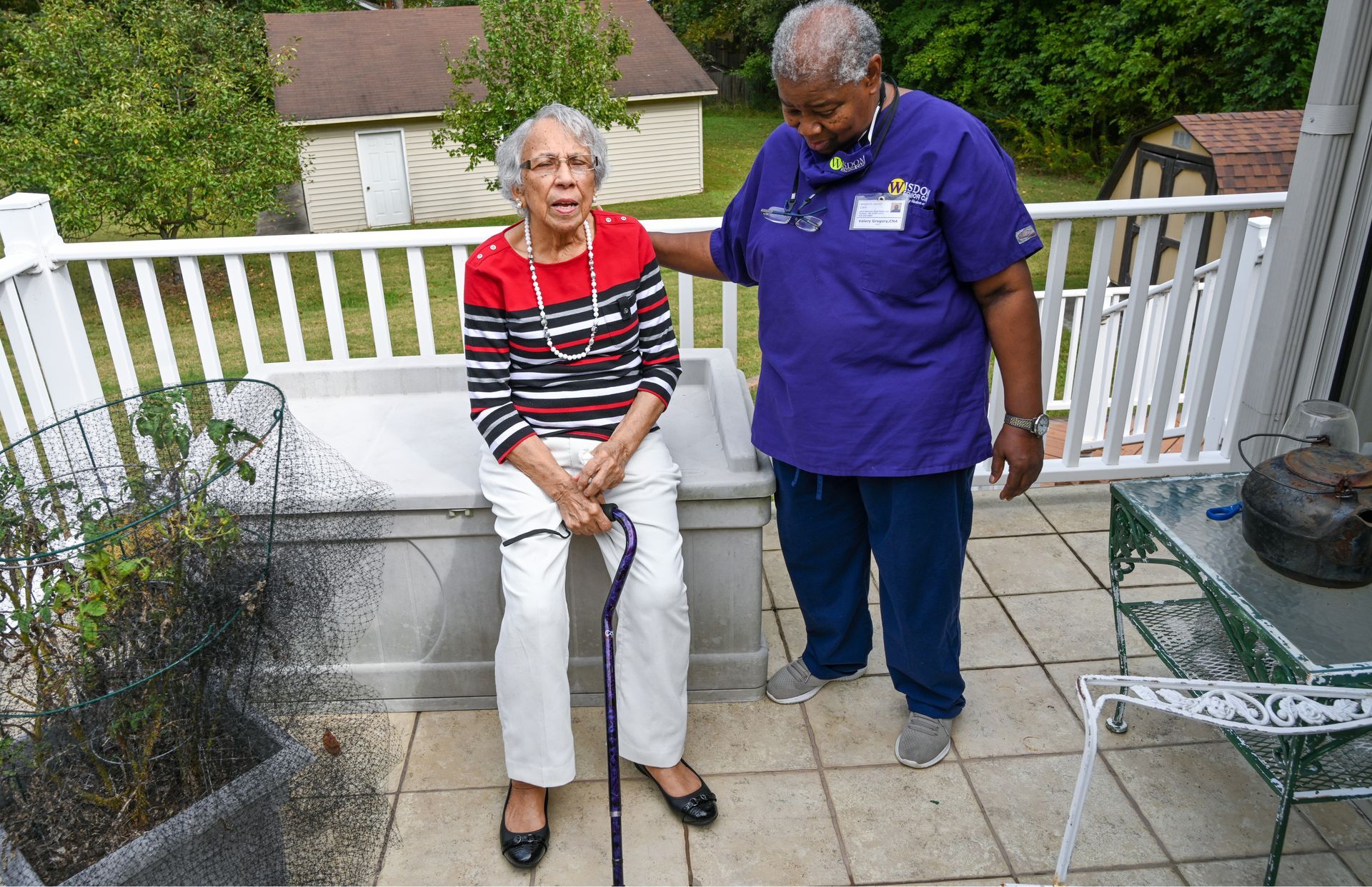 Woman with cane assisted by caregiver on a porch; caregiver's hand on her back for support.