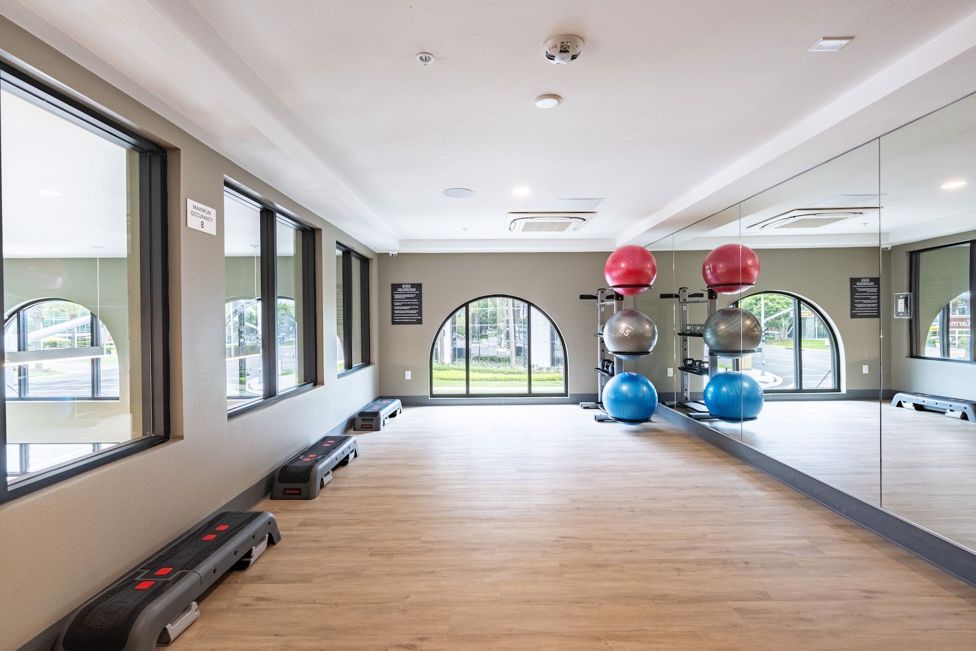 Gym interior with exercise balls, mirrors, windows, and step platforms. Wooden floor and neutral colored walls.