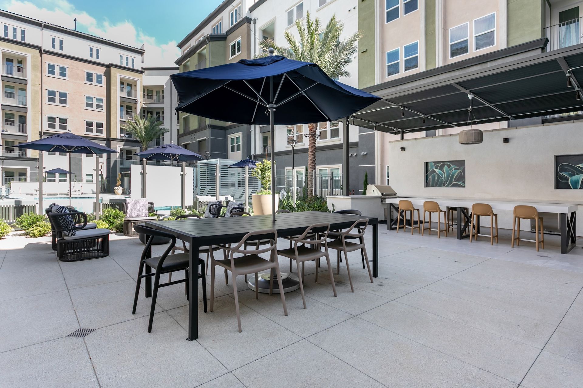 Outdoor patio with tables, chairs, umbrellas, and a grilling area in an apartment complex courtyard.