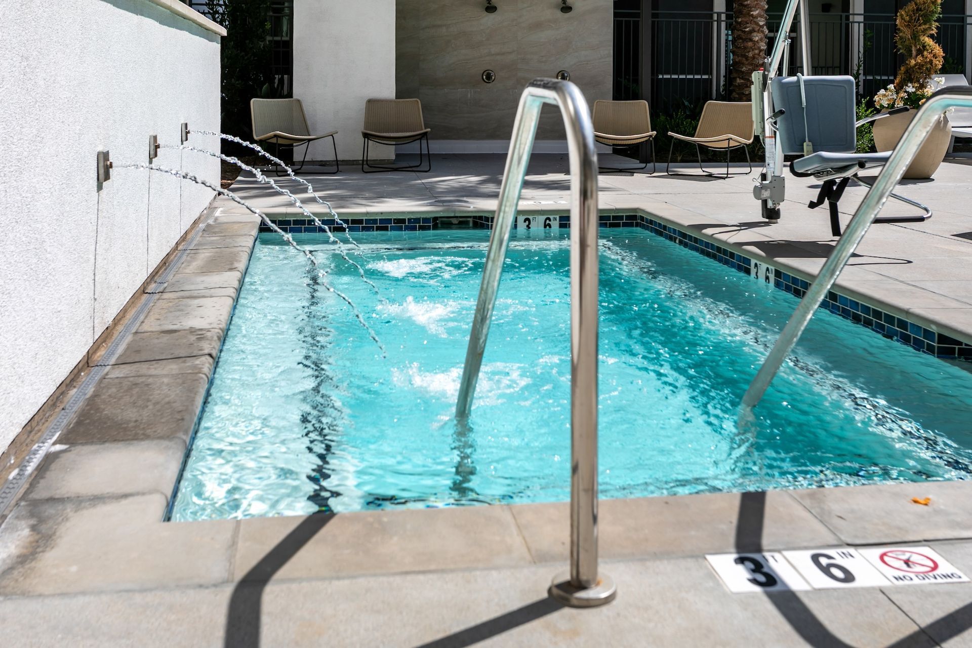 A small rectangular spa pool with bubbling turquoise water, metal handrails, and jets spraying water; patio seating in background.