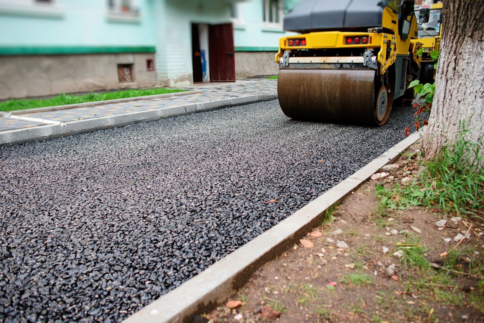 Road roller compacts fresh asphalt along a curb on a residential street.
