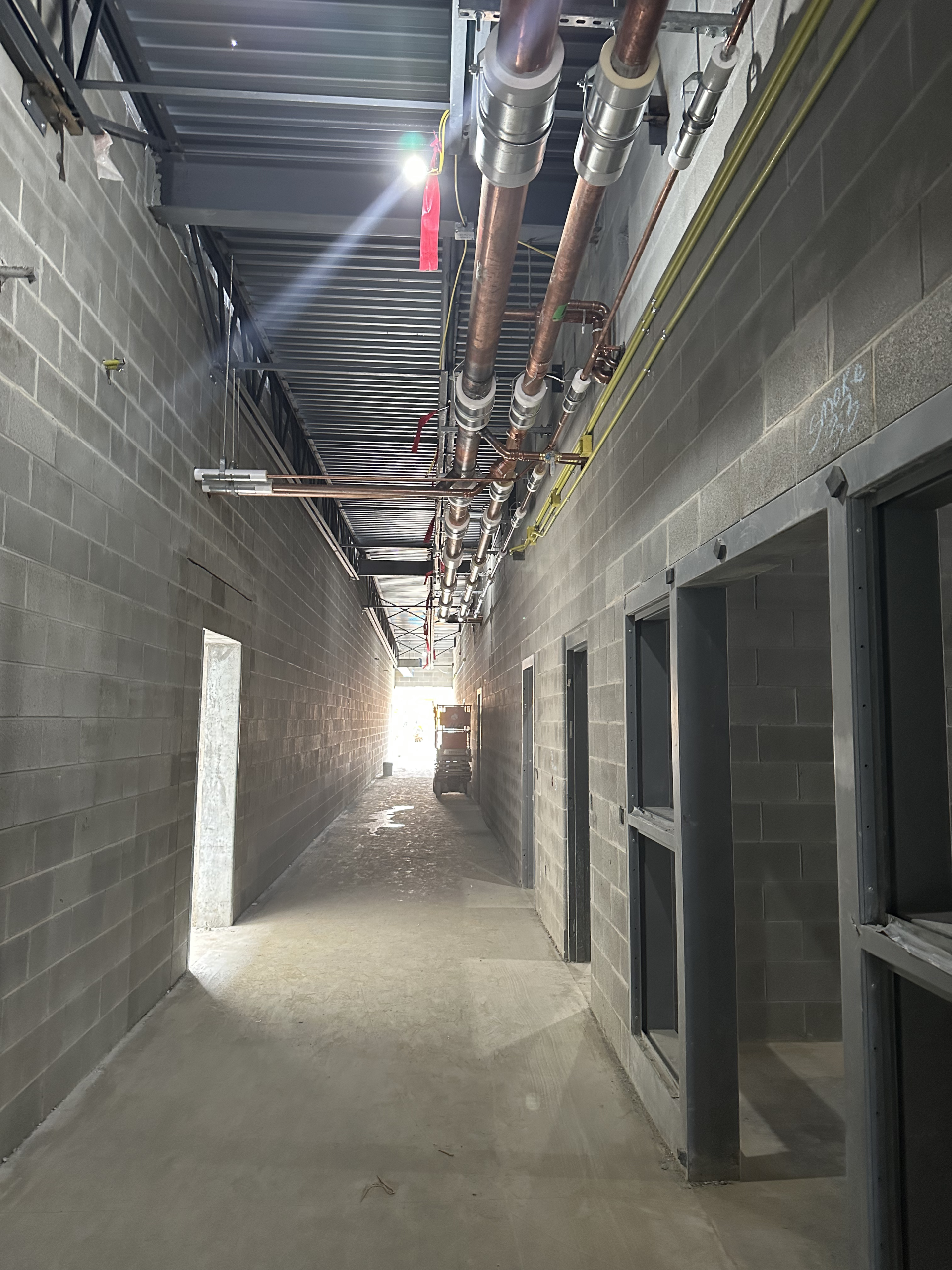 Interior view of a building under construction; a long hallway with unfinished walls, doors, and overhead pipes.