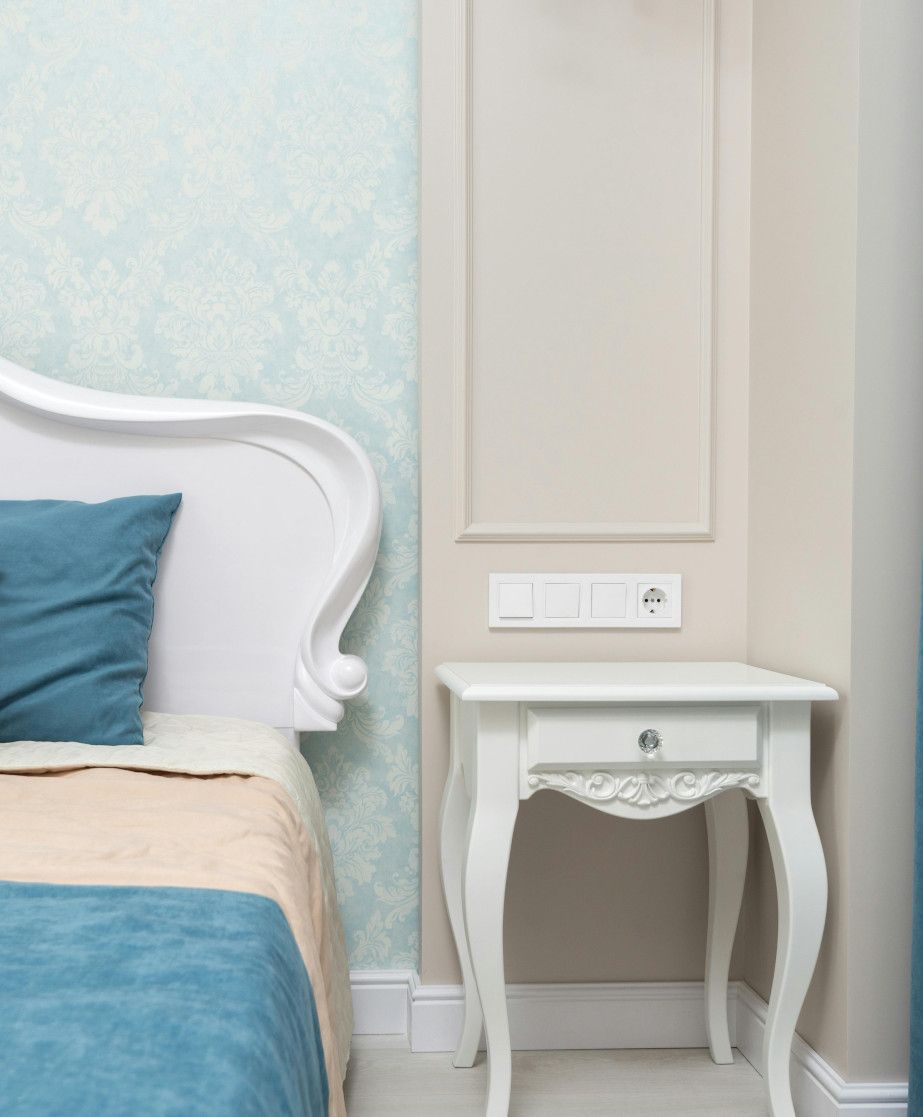 White bedside table with drawer and ornate legs next to a bed with a teal bedspread.