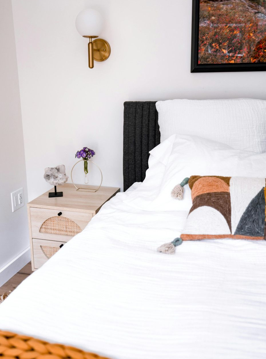 Bedroom with white linens, decorative pillow, gold sconce, and wooden nightstand.