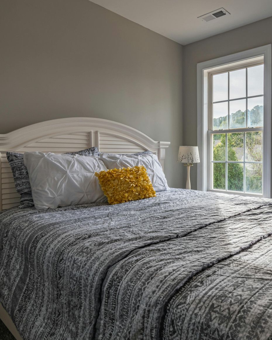 Bedroom with a white headboard, gray bedding, a yellow pillow, and a window with a view of trees.