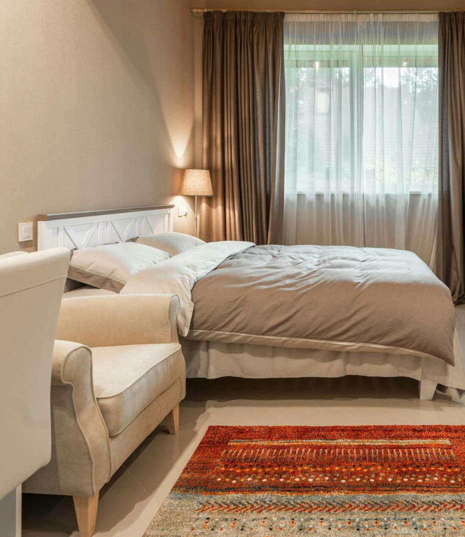 Bedroom with a bed, armchair, rug, and curtains. Earth tones dominate with a light-filled window.