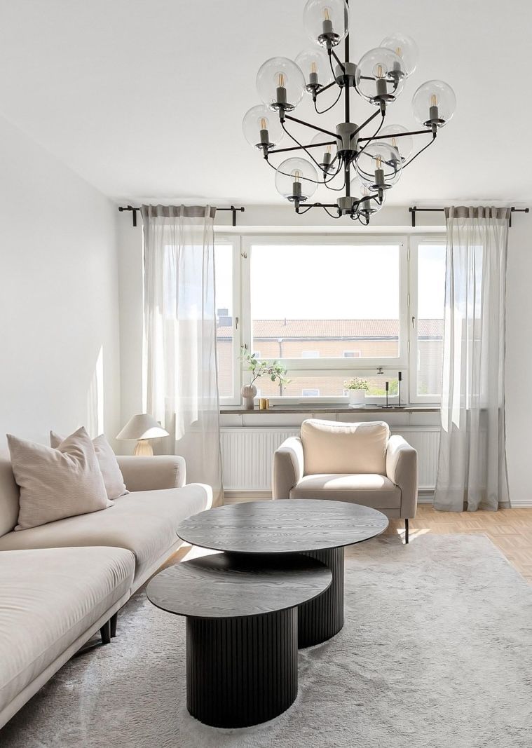 Living room with beige sofa, armchair, and coffee tables under a chandelier, next to a sunny window.