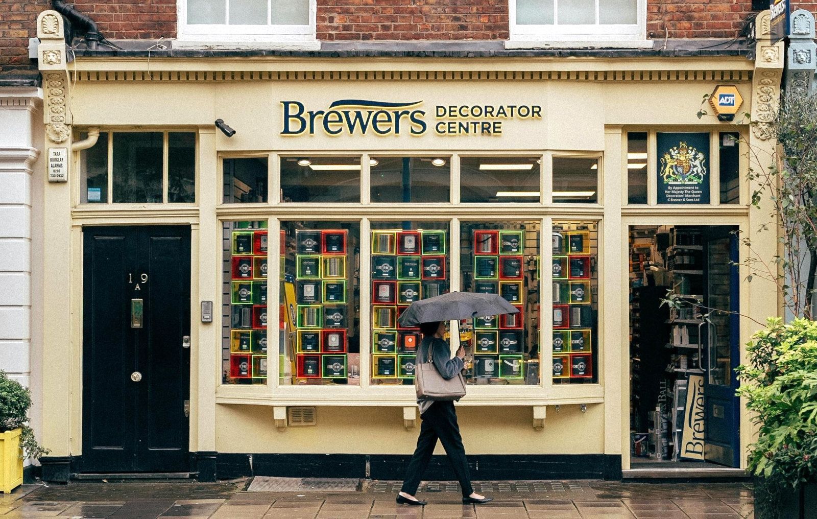 A person is walking in front of a brewers decorator centre store holding an umbrella.