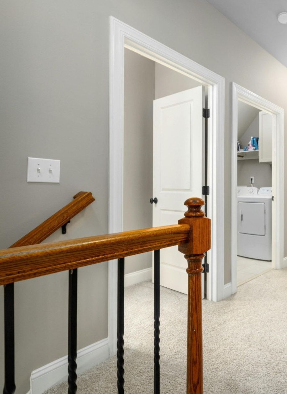 Hallway with staircase, open door, and laundry room. Gray walls, carpet. Oak banister, white doors.