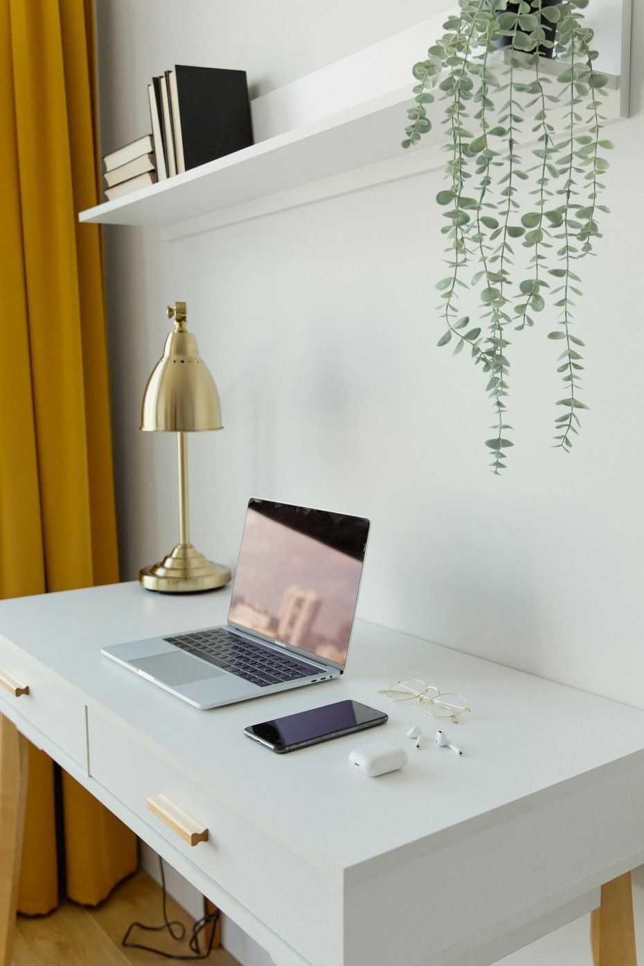 White desk with laptop, phone, lamp, and hanging plant. Yellow curtain in the background.
