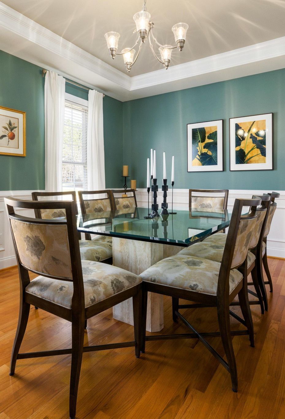 Dining room with a glass table, chairs, teal walls, chandelier, and artwork.