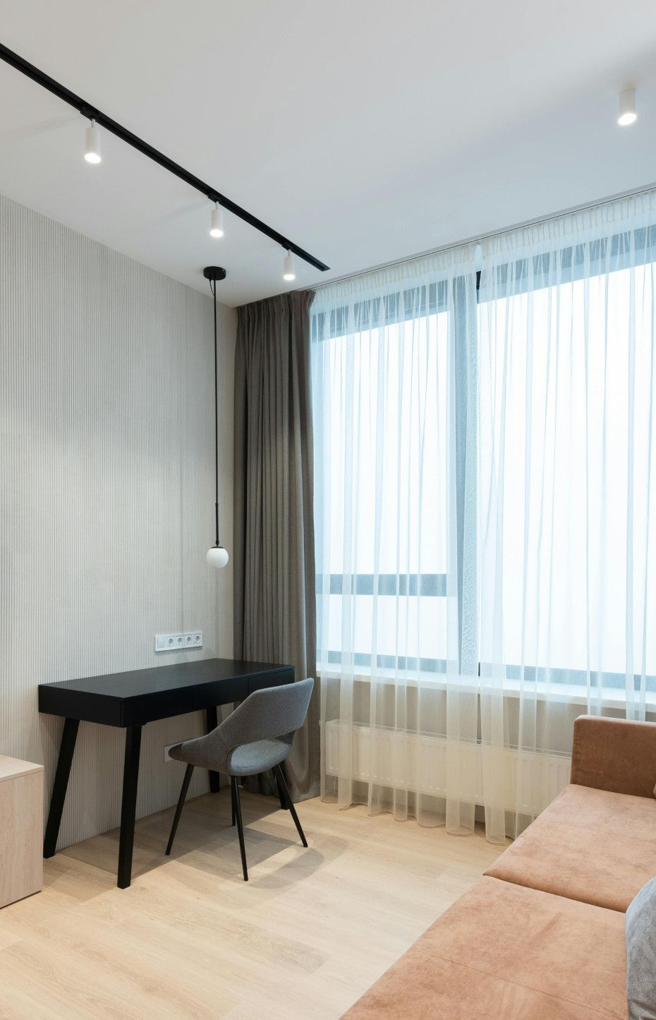 A modern room with a desk, chair, and sofa near a window with sheer curtains and wood flooring.