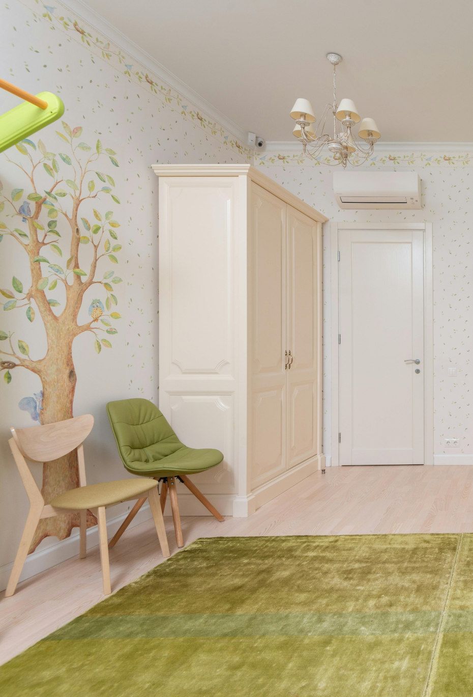 Child's bedroom with green rug, two chairs, cream wardrobe, tree mural, and white door.