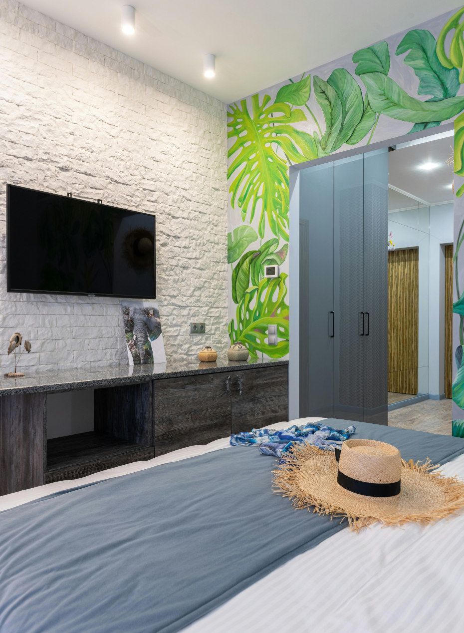 Bedroom with brick wall, TV, built-in storage, and tropical mural. A hat rests on a blue bedspread.